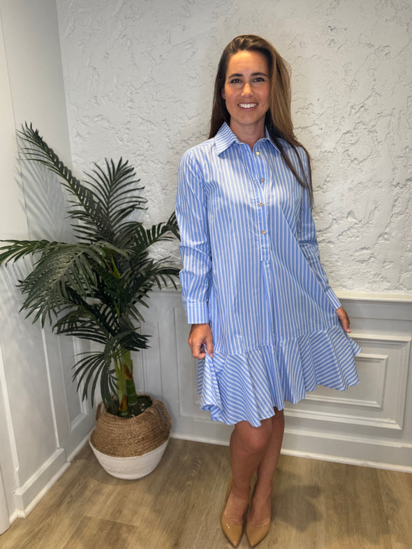 Smiling model in front of a white wall and a tropical plant in the corner wearing the Duffield Lane Iris Drop Waist Shirt Dress - Light Blue Double Stripe. A mini/knee length, long sleeve, seersucker dress. It has a collar and buttons down the front and a ruffle at the bottom hem. 
