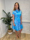 Smiling model in front of a white wall with a plant in the corner, in the Patty Kim Colette Dress - Aqua Rococo. A short sleeved mini dress with a drop waist and buttons down the front. A white and royal blue floral pattern on an aqua background