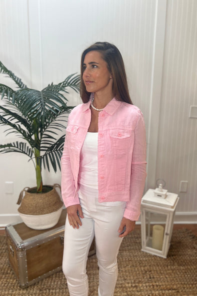 A woman stands angled wearing a light pink linen jacket with a white tank topand white pants with plant and antique trunk in the background