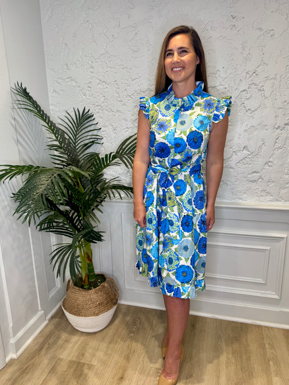 Smiling model in front of a white wall with a plant in a corner wearing the Patty Kim Megan Dress - Blue Zinnias. A midi length flutter sleeve and collar, with a tie waist and a ruffle at the bottom hem, with blue and green flower pattern
