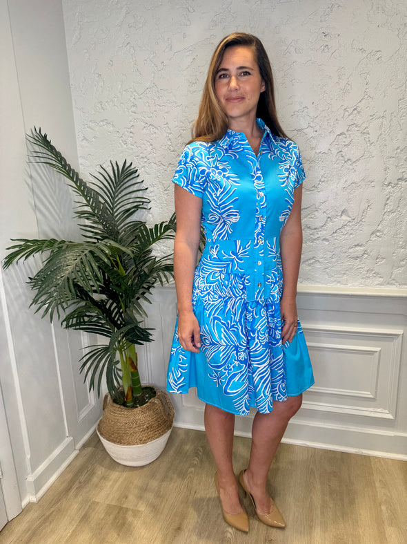 Smiling model in front of a white wall with a plant in the corner, in the Patty Kim Colette Dress - Aqua Rococo. A short sleeved mini dress with a drop waist and buttons down the front. A white and royal blue floral pattern on an aqua background