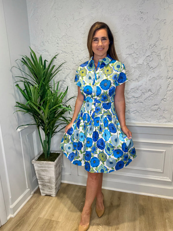 Model in front of a white wall in the Patty Kim Collette Dress - Blue Zinnias. A knee length dress with short sleeves and a drop waist and buttons down the front