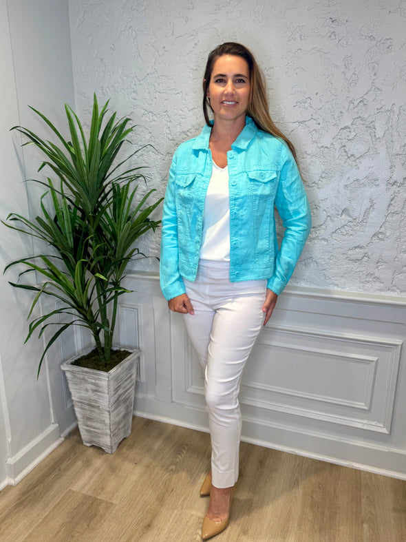 Smiling model in front of a white wall with a green plant in the corner. She's wearing the Lulu B Linen Travel Jacket - Seafoam, a light linen jacket with 2 front pockets and lightly frayed hems. Paired with a white shirt and white pants. 
