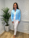 Smiling model in front of a white wall with a green plant in the corner. She's wearing the Lulu B Linen Travel Jacket - Clear Blue, a light linen jacket with 2 front pockets and lightly frayed hems. Paired with a white shirt and white pants. 
