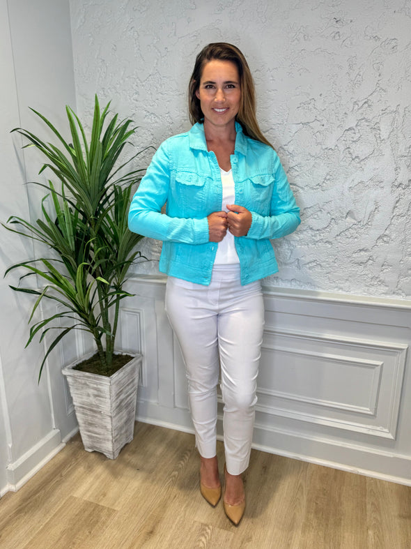 Smiling model in front of a white wall with a green plant in the corner. She's wearing the Lulu B Linen Travel Jacket - Seafoam, a light linen jacket with 2 front pockets and lightly frayed hems. Paired with a white shirt and white pants. 