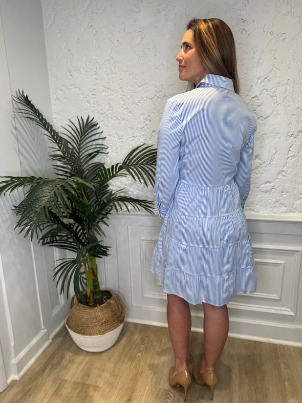 Back view of model in front of a white wall wearing the Patty Kim Capri Dress - Blue/White Stripe. A long sleeve, knee length dress that is tiered and has gold buttons down the front, with a collar. 