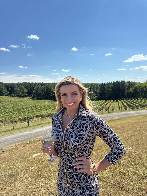 Woman in a black and beige pattern and a tie detail on the front, in a vineyard with a glass of wine