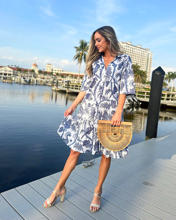 Model on a dock wearing a navy and white, knee length dress, with a floral and bird print, and holding a bamboo bag