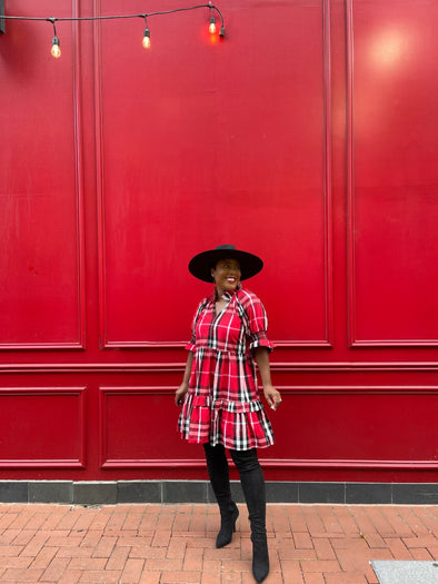 Model wearing Duffield Lane Skye Dress in Red Plaid with black hat against bright red wall