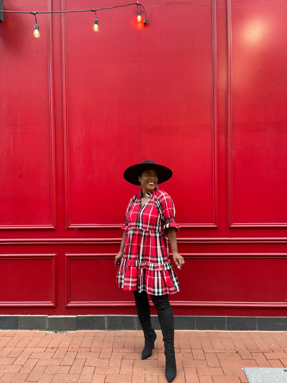 Model wearing Duffield Lane Skye Dress in Red Plaid with black hat against bright red wall