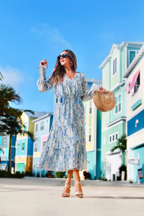 Model in a tiered midi dress with a ruched waist, sheer long sleeves, and v neck. It has a beautiful blue, purple, metallic gold floral print, that is both subtle and stunning