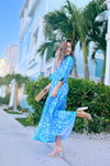 Model in a blue/ aqua patterned tiered maxi dress, with touches of green and white and other shades of blue, kicking her leg up.