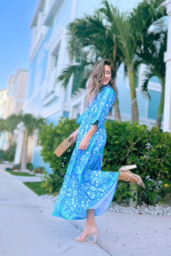 Model in a blue/ aqua patterned tiered maxi dress, with touches of green and white and other shades of blue, kicking her leg up.