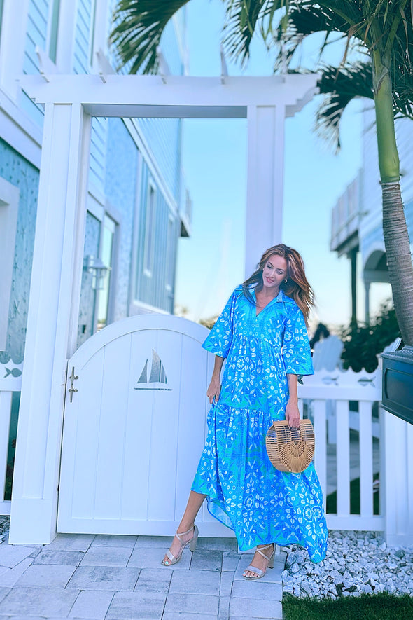 Model in front of a gate in a blue/ aqua patterned tiered maxi dress, with touches of green and white and other shades of blue. 