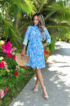 Woman wearing a blue knee-length shirt dress with a white tropical print, tie waist, and rolled sleeves with a green contrast cuff. 