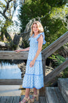 Woman in a blue dress standing on a wooden bridge by a body of water with trees in the background.