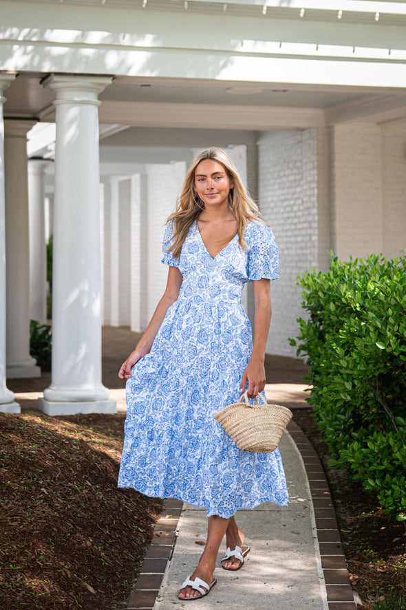 Woman in a blue floral dress holding a straw bag on a sunlit path.