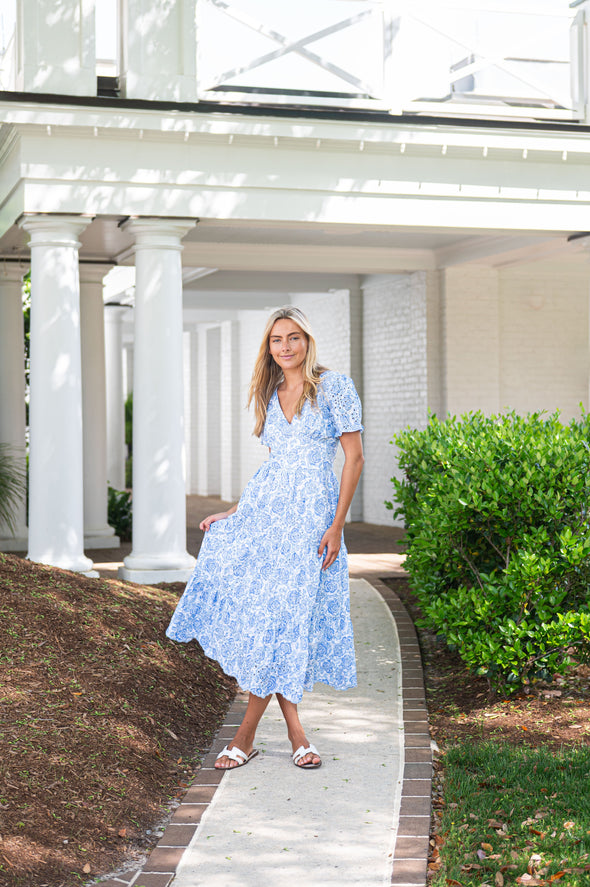 Woman in a blue dress standing on a pathway with white columns and greenery in the background