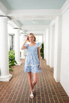 Woman in a blue floral dress standing in a sunlit hallway with white columns and plants.