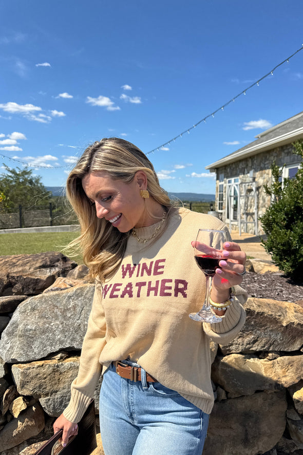 Person wearing a 'Wine Weather' sweatshirt holding a glass outdoors.