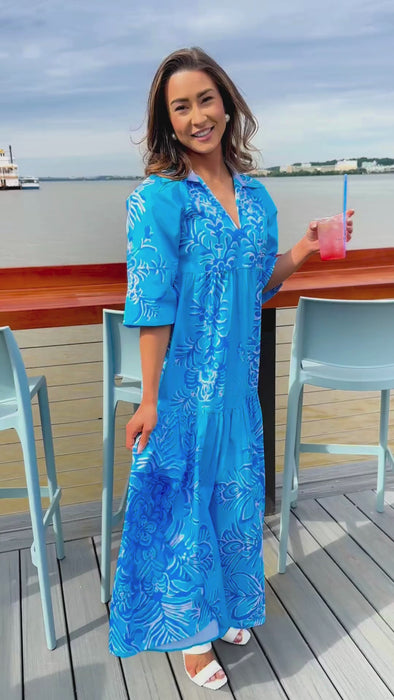 Woman wearing a bright blue maxi dress with white floral pattern. The dress has a V-neck, flared sleeves, and a tiered skirt. She’s wearing white sandals and standing against a plain white background.