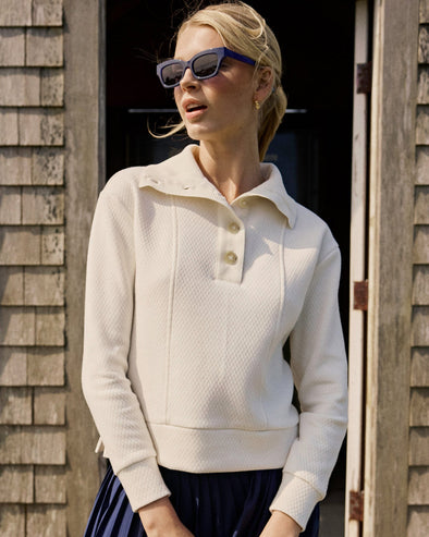 Woman wearing a cream-colored sweater with a collar, standing against a wooden wall.