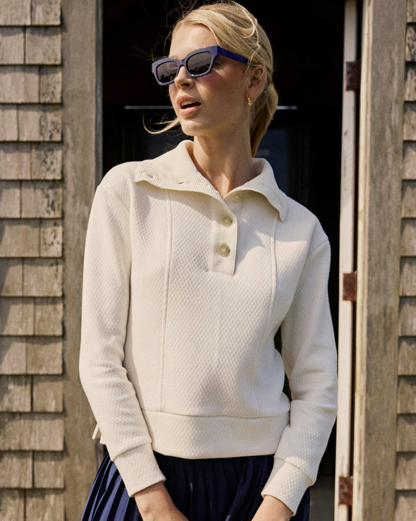 Woman wearing a cream-colored sweater with a collar, standing against a wooden wall.